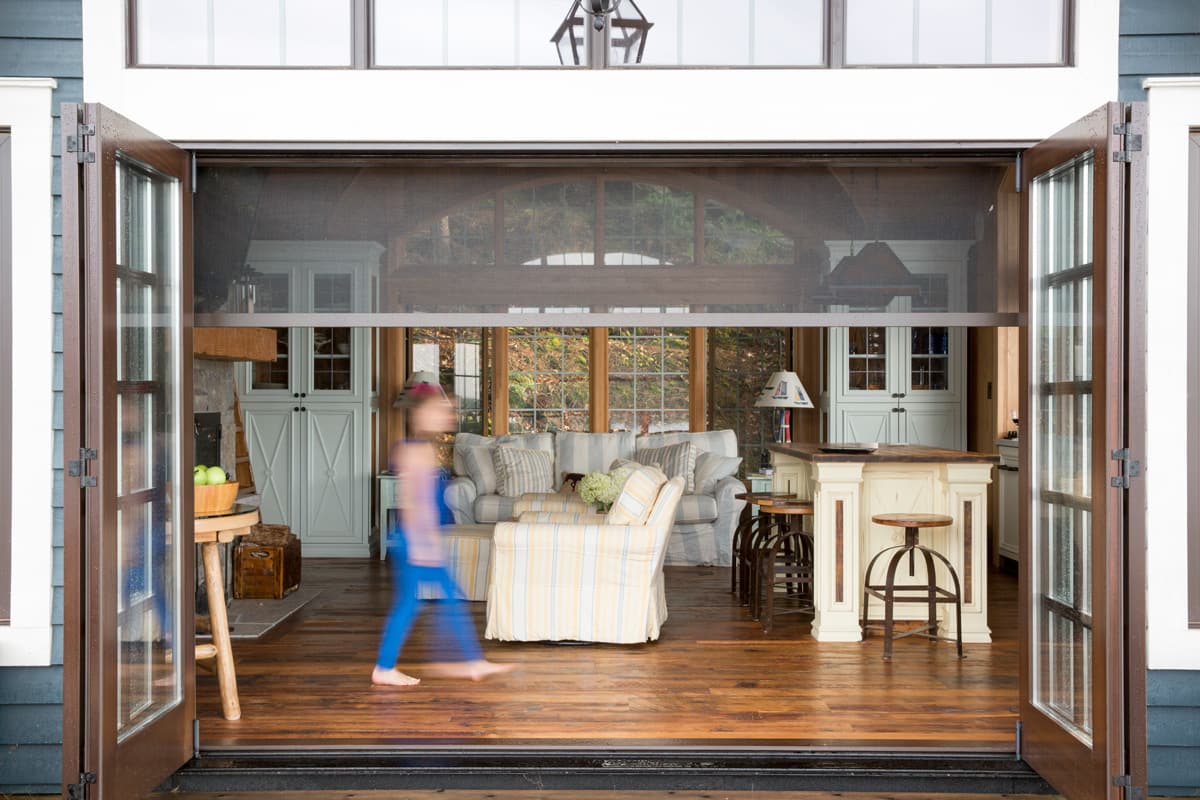 The image is of a person walking past the open sliding doors of a cozy living room with wooden floors, a plaid sofa, and a kitchen area with a bar stool and lamp, demonstrating automation systems, and automated window coverings.